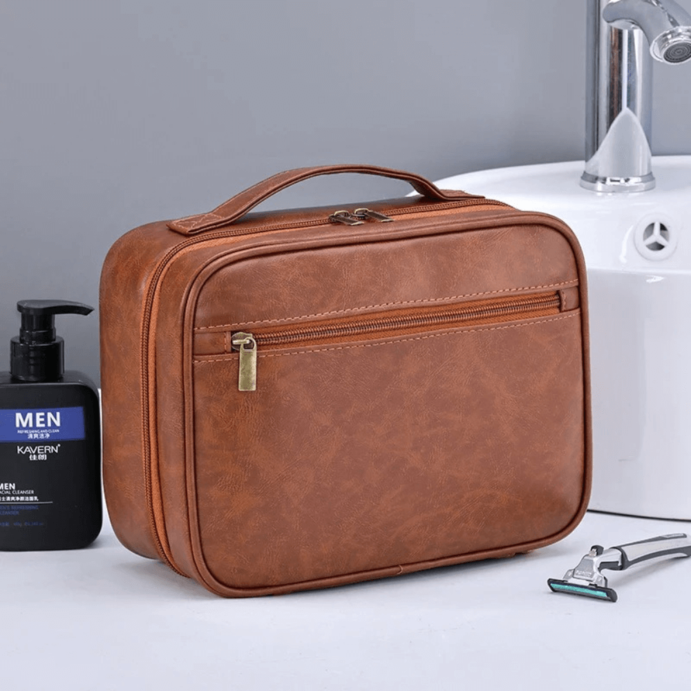 Brown toiletry bag on a bathroom counter with a sink and shaving tools in the background.